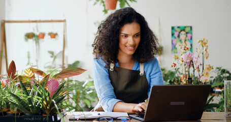 Young Afro American woman flower business owner in apron using laptop working at the floral store they checking the inventory. Floristry and modern technology concept. Slow motion.