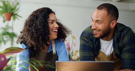 Close up. African American young couple florists in aprons working in flower store and talking typing on laptop. Woman working at laptop together with man florist using an online service to accepting