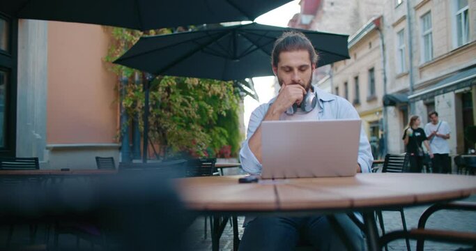 Young male businessman in casual clothes working online at freelance. Attractive bearded man sale manager using laptop for work while sitting at cafe shop outdoors on urban street.