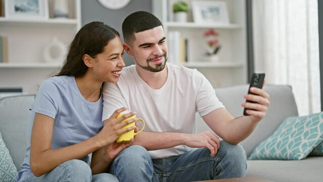 Beautiful couple enjoying their breakfast coffee together, sitting casually on their living room sofa, engrossed in a lively online conversation via mobile video call at home