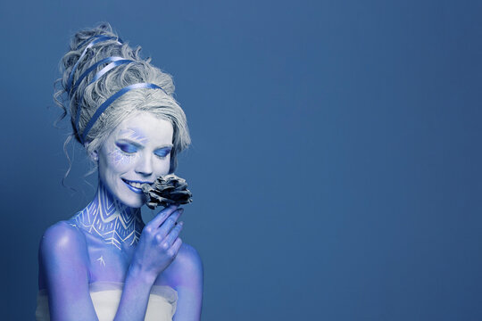 Happy laughing woman fashion model with blue skin, stage makeup and hairstyle holds black rose flower on blue background
