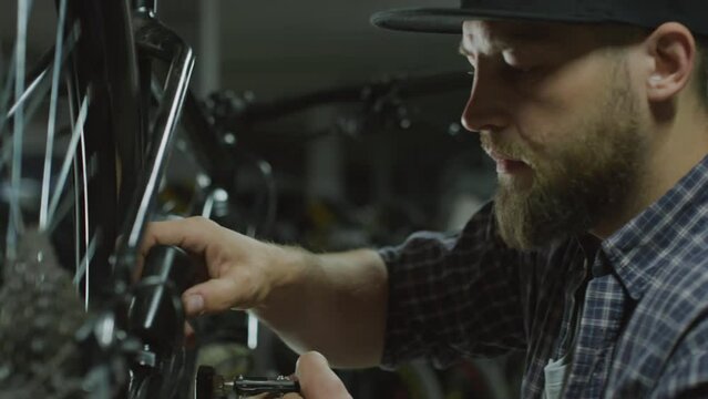 Handheld shot of man cranking bike pedal and applying lubricant to chain while working in repair shop