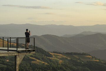 A man stands on a lookout and takes a photo of the mountain landscape with his smartphone