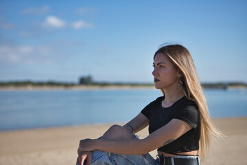 Fototapeta na wymiar Portrait of young, beautiful blonde woman, green eyes, with black top, torn jeans and tattoos, sitting, looking at infinity with the sea in the background. Concept of peace, tranquility, relaxation.