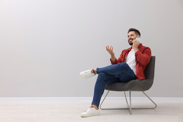 Handsome man talking on smartphone while sitting in armchair near light grey wall indoors, space for text