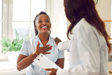 Happy female patient thanking her doctor from the bottom of her heart. Joyful, smiling, grateful, relieved, young African American woman holding hand on her chest and saying Thank you to physician - obrazy, fototapety, plakaty