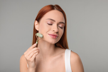 Young woman massaging her face with jade roller on grey background