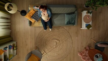 In the shot from above, a pregnant woman sits on a sofa in a room and holds her head. She has a...