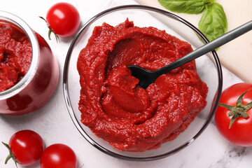 Glass bowl and jar of tasty tomato paste with spoon and ingredients on white marble table, flat lay