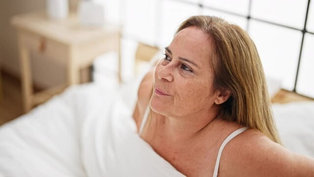 Middle age hispanic woman waking up sitting on bed at bedroom