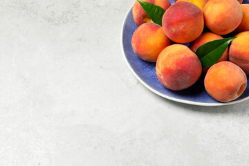 Many whole fresh ripe peaches and green leaves in plate on white table, space for text