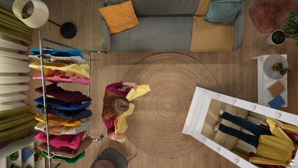 Top view of living room. Girl holding sweatshirt with yellow shirt, trying it looking at herself in the mirror, hanger with clothes next to her.