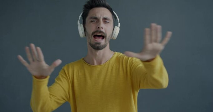 Slow motion portrait of joyful young man wearing wireless headphones dancing listening to music having fun on grey color background