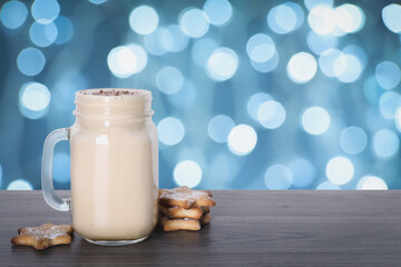 Delicious eggnog in mason jar and cookies on wooden table against blurred Christmas lights, space for text