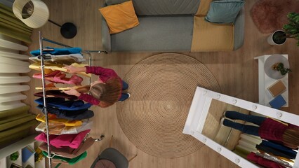 Top view of living room. Girl standing next to hanger with clothes and looking through many colorful clothes on it, picking outfit.