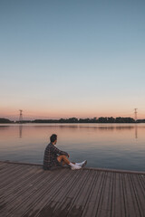 Young 25 year old brown man with plaid shirt sitting on the end of a wooden pier during sunset, watching the calm water surface in Almere Netherlands