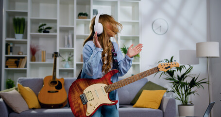 Energetic funny girl with long hair playing electric guitar, sing song and dancing at living room...