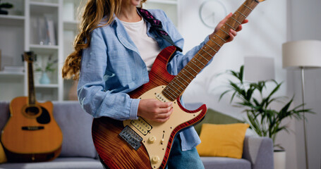Close up of long haired young woman rocker in denim shirt playing acoustic guitar, singing and dancing at light living room at home - obrazy, fototapety, plakaty