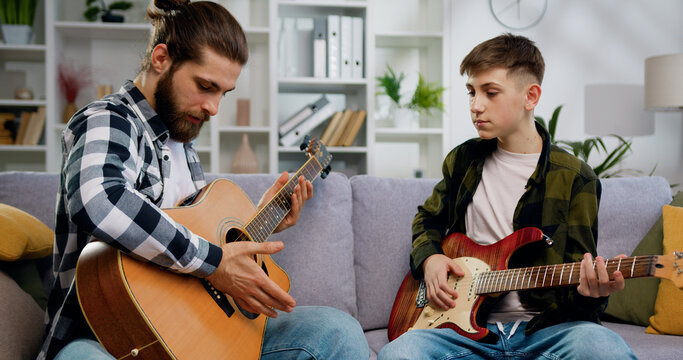 Talented music teacher and teenage student boy sitting on sofa learning to play on guitar in private lesson in light living room or music studio.