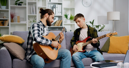Talented music teacher and teenage student sitting on sofa playing guitar together in private...