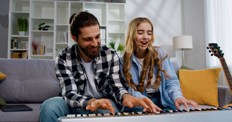 Funny portrait of smiling father with daughter playing synthesizer singing dancing and sitting sofa at light living room. Activity family love and lifestyle Concept - obrazy, fototapety, plakaty