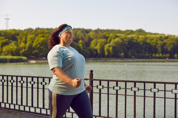 Body positive plus size woman jogging outdoors and having sport walking losing weight. Happy smiling fat girl having sport training in nature walking on the bridge along the river in the city park. - obrazy, fototapety, plakaty