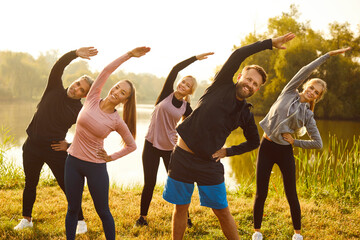 Group of people having an outdoor fitness workout. Five happy men and women in activewear doing sports exercises in nature. Smiling male and female athletes standing on a river bank doing a side bend - obrazy, fototapety, plakaty