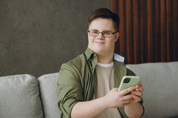 Young happy man with down syndrome wear glasses casual clothes use mobile cell phone sits on grey...