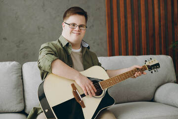 Young fun man with down syndrome wear glasses casual clothes play guitar look camera sits on grey...