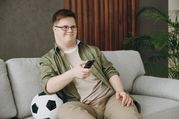 Young man with down syndrome wear glasses casual clothes watch tv football live stream sits on grey...