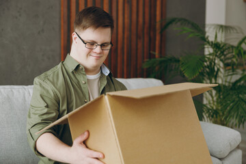 Young man with down syndrome wear glasses casual clothes unpacking empty cardboard box sits on grey...