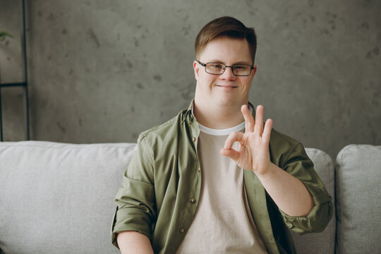 Young smiling man with down syndrome wear glasses casual clothes show ok okay gesture sits on grey sofa couch stay at home flat rest spend free time in living room. Genetic disease world day concept. - Powered by Adobe