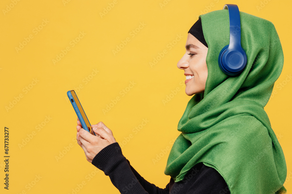 Wall mural side view young muslim woman wear green hijab abaya black clothes headphones listen music use mobile