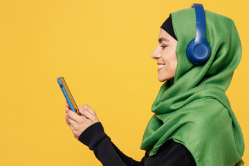 Side view young muslim woman wear green hijab abaya black clothes headphones listen music use...