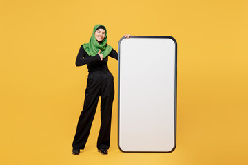 Full body young asian muslim woman wear green hijab abaya black clothes big huge blank screen mobile cell phone show thumb up isolated on plain yellow background. People uae islam religious concept.
