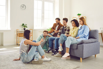 Group of friends hanging out together. Several young people meeting at somebody's place. Young men and women sitting on sofa and listening to girl who is sitting on floor and talking about something