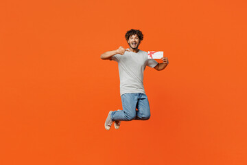 Full body young smiling happy Indian man he wears t-shirt casual clothes jump high hold point finger on gift certificate coupon voucher card for store isolated on orange red color background studio.