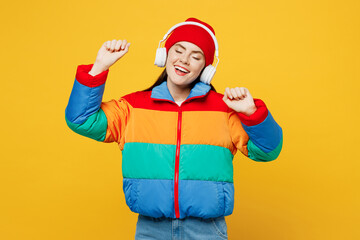 Young smiling happy woman she wears padded windbreaker jacket red hat casual clothes listen to music in headphones raise up hands dance isolated on plain yellow background studio. Lifestyle concept.