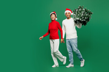 Full body side view merry young couple man woman wearing red casual clothes Santa hat posing hold Christmas walk go look camera isolated on plain green background. Happy New Year 2024 holiday concept.