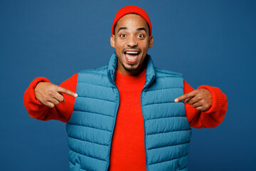 Young surprised shocked man of African American ethnicity wear padded vest red hat point index...