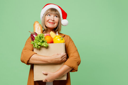 Elderly happy woman wear brown shirt casual clothes red Santa hat hold shopping paper bag with food products look aside ona rea mockup isolated on plain green background. Delivery service from shop.