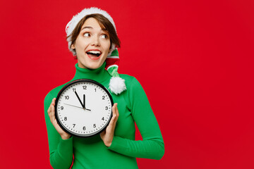 Young smiling cheerful woman wear green turtleneck Santa hat posing hold in hand clock look aside...