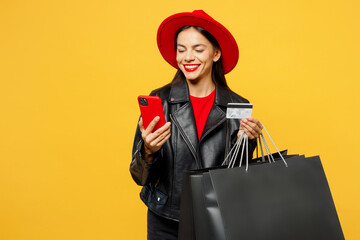 Young minded pensive woman wears casual clothes red hat hold shopping paper package bags use mobile cell phone credit bank card isolated on plain yellow background. Black Friday sale buy day concept. - obrazy, fototapety, plakaty