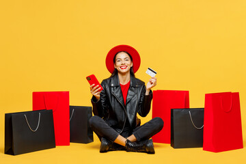 Full body young woman wear casual clothes red hat sit near shopping paper package bags hold mobile cell phone credit bank card isolated on plain yellow background. Black Friday sale buy day concept.