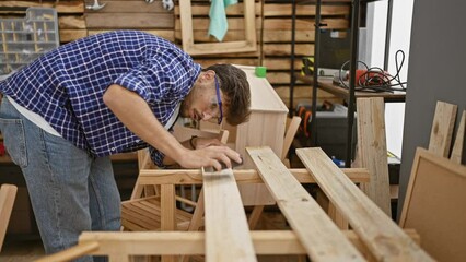 Handsome, serious, young arab man working diligently, sanding wood plank at the heart of his busy, indoor carpentry workshop