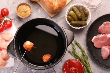 Fondue pot, forks with fried meat pieces, pickled cucumbers and other products on grey textured table, flat lay