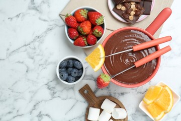 Fondue pot with melted chocolate, marshmallows, fresh orange, different berries and forks on white marble table, flat lay. Space for text