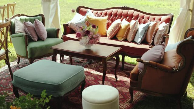 A decorated area for guests to relax at the wedding, a wooden table and a beautiful bouquet on it, pouf and a red antique sofa with pillow on it, slow motion