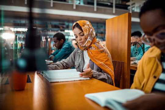 Focused young woman in vibrant hijab immersed in her studies at a bustling library