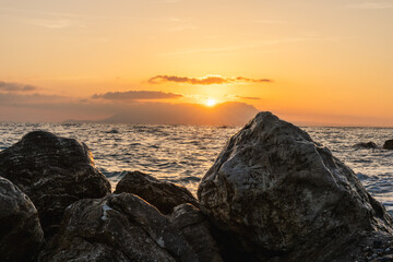 Sunrise over the ocean. The sun is a golden orange color partially obscured by clouds. In the...